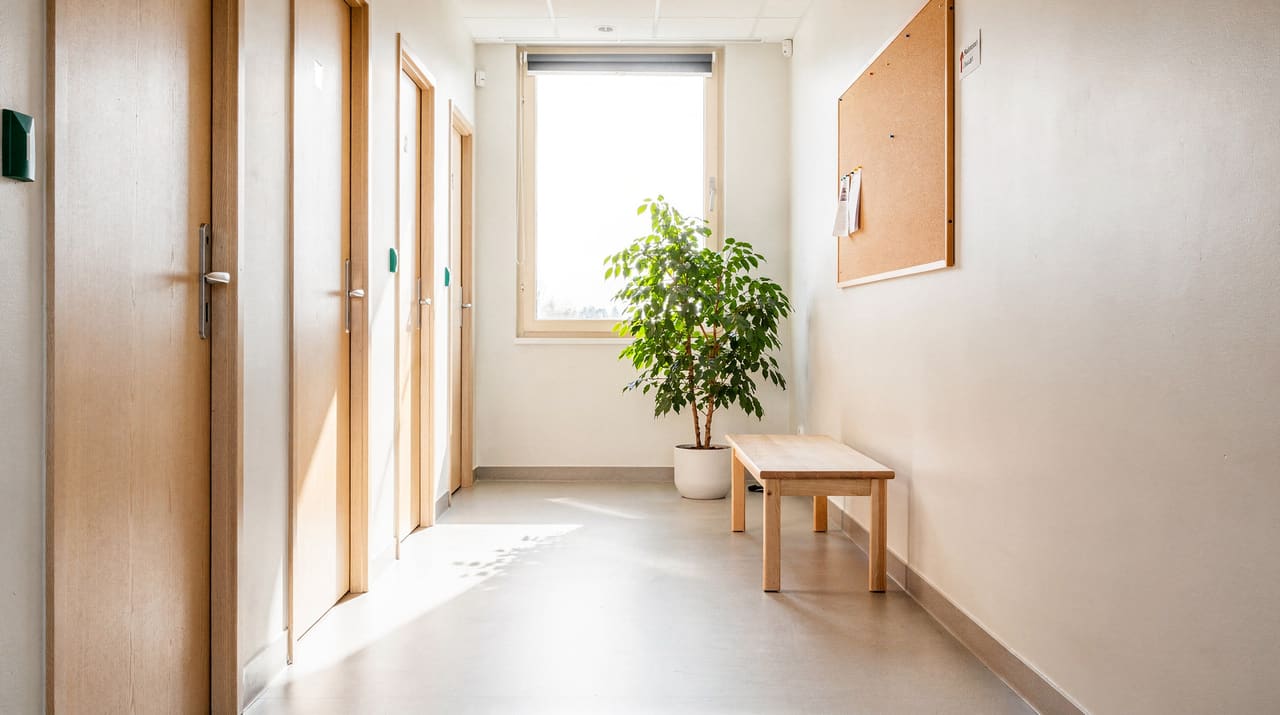 En ljus korridor i ett svenskt vård- och boendehus i morgonljus med en rad dörrar, en bänk, en grönväxt och ett stort fönster i fjärran