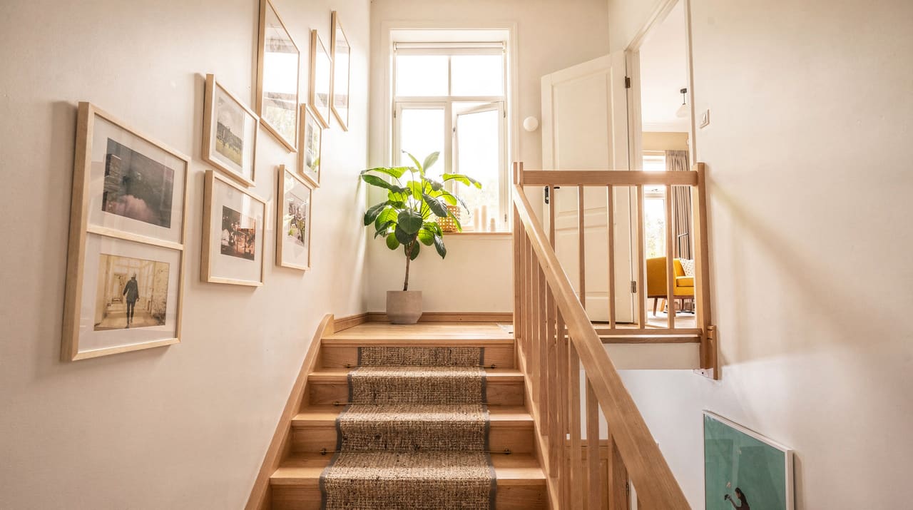 Ett ljust svenskt trapphus med vilplan i morgonljus, träräcke, en löpare i trappan, inramade tavlor på väggen och en grönväxt vid ett fönster