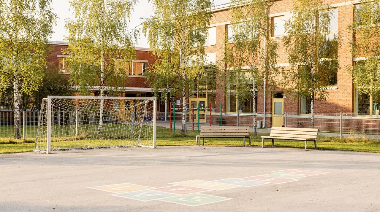 En solig skolgård i september med hopphage målad på asfalten, ett fotbollsmål, björkar längs staketet och en tegelskola i bakgrunden